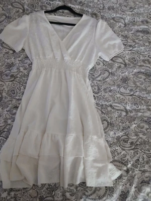 Vit omlottklänning med volang - Söt vit kortklänning med omlott och v-ringning. Klänningen har korta ärmar, smock i midjan och volangdetaljer nedtill. Tillverkad i ett lätt tyg med diskret prickigt mönster som ger en fräsch och somrig känsla. Kan skicka fler bilder vid förfrågan 