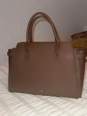 Brun handväska Anna Field - Snygg brun handväska från Anna Field i stilren design med två handtag och guldfärgade metalldetaljer. Sparsamt använt och har en längre axelrem som man kan koppla till väskan. Rymmer dator och flera böcker så passar perfekt till skolan.