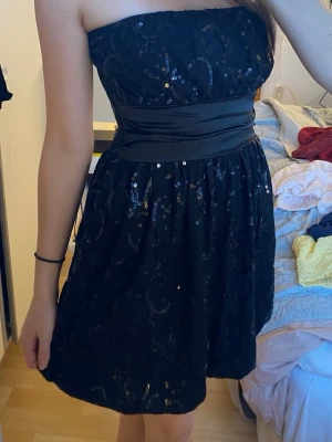 Svart paljettklänning med markerad midja - bandeu klänning  - Snygg svart axelbandslös klänning med paljetter och markerad midja i satinliknande material. Kort modell som passar perfekt när du vill sticka ut lite extra. Klänningen har en glittrig yta och en feminin siluett. Storlek S/M men mer som en S