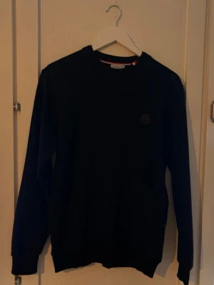 Svart sweatshirt från Moncler - En stilren svart sweatshirt från Moncler med rund halsringning och diskret Moncler-logga på bröstet. Tröjan har långa ärmar och ribbade muddar vid ärmslut och nederkant. Perfekt för en clean och avslappnad look.