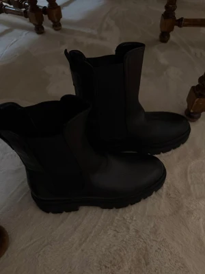 Svarta Chelsea boots från Tamaris - Säljer ett par svarta Chelsea boots från Tamaris med chunky sula och högt skaft. Skorna är i slätt läder med elastiska paneler på sidorna och dragflikar både fram och bak. Perfekta för en stilren och edgy look.