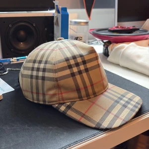 Burberry keps - Säljer en klassisk beige keps från Burberry med det ikoniska rutmönstret i svart, vitt och rött. Kepsen har böjd skärm och justerbar snapback baktill. Märkt med 'BURBERRY LONDON ENGLAND' på baksidan. Perfekt accessoar för en trendig streetwear-look.