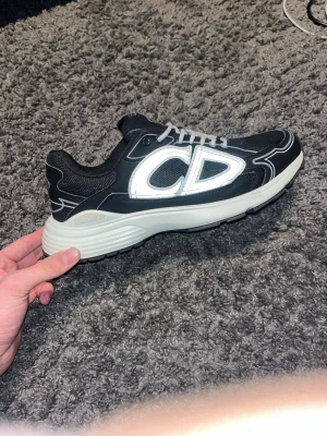 Svarta Dior sneakers med vita detaljer - Snygga svarta sneakers från Dior med vita kontrastdetaljer och stora CD-loggan på sidan. Skorna har mesh och lädermaterial, vita snören och en tjock vit sula som ger en sportig look. Perfekta för dig som vill sticka ut med exklusiva sneakers.
