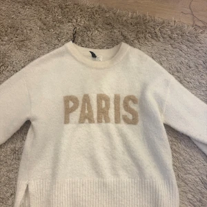 Vit stickad tröja med PARIS-tryck - Säljer en vit stickad tröja med beige texten PARIS framtill. Tröjan har rund halsringning, långa ärmar och ribbade detaljer vid ärmslut och nederkant. Perfekt för dig som gillar enkel men trendig stil.
