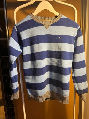 Blå randig stickad tröja Tommy Hilfiger - Snygg stickad tröja från Tommy Hilfiger med breda blå och ljusblå ränder. Tröjan har ribbstickade muddar och krage i beige, samt ett litet logomärke på bröstet. Perfekt för dig som gillar klassisk och avslappnad stil.