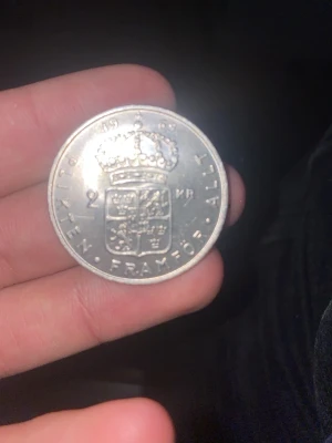 Svenskt 2-kronorsmynt 1956 silver - Säljer ett svenskt 2-kronorsmynt från 1969 i silverfärg med Gustaf VI Adolfs porträtt på ena sidan och kunglig krona samt riksvapnet på andra sidan. Texten 'PLIKTEN FRAMFÖR ALLT' och 'SVERIGES KONUNG' finns på myntet. Perfekt för dig som gillar samlarobjekt och svensk historia. 13 gram silver