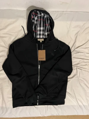 Svart hoodie från Burberry med dragkedja - Snygg svart hoodie från Burberry i storlek XXL men passar som en L med dragkedja och klassiskt rutigt foder i huvan. Märkets logga är diskret broderad på bröstet. Hoodien har dragsko i huvan och ribbade muddar. Perfekt för en stilren och avslappnad look.