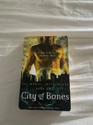 City of Bones av Cassandra Clare - Säljer boken 'City of Bones' av Cassandra Clare, första delen i serien The Mortal Instruments. Omslaget är färgstarkt med en stadssilhuett och en tatuerad överkropp i gult och svart. Pocketbok på engelska, perfekt för dig som gillar urban fantasy och äventyr.