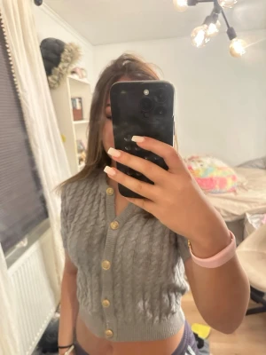 Grå kabelstickad kort kofta - Säljer en grå kabelstickad kofta med kort passform och stora beige knappar framtill. Koftan har v-ringning och ribbade kanter. Perfekt för att skapa en trendig lager-på-lager look.