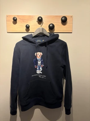 Mörkblå hoodie Polo Ralph Lauren - Mörkblå hoodie från Polo Ralph Lauren med det ikoniska björn trycket på. Storleken är M men sitter mer som S. Sitter väldigt bra och är otroligt skön. Perfekt för dig som vill ha en avslappnad hoodie som man kan ha när som helst. Priset är inte hugget i sten så tveka inte att lämna prisförslag!