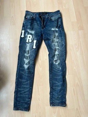 Amiri Jeans - Säljer ett par blå jeans med slitningar och stora vita patchbokstäver på både fram- och baksidan. Jeansen har klassisk femficksdesign och raka ben. Materialet är denim och de har en cool, trendig look med tydliga slitningar och broderade detaljer.