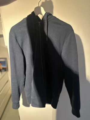 Mörkblå zip-hoodie med huva jack and jones  - En enkel och stilren mörkblå hoodie med dragkedja och huva. Tröjan har långa ärmar, ribbade muddar och snören i huvan. Perfekt för att slänga på sig när det är lite kyligt. Materialet känns mjukt och bekvämt.