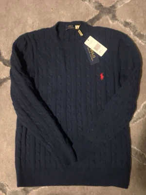 Mörkblå kabelstickad tröja Polo Ralph Lauren - Mörkblå kabelstickad tröja från Polo Ralph Lauren med rund halsringning och klassisk röd broderad logga på bröstet. Tröjan har långa ärmar och ribbade muddar vid ärmslut och nederkant. Perfekt för dig som gillar stilren och tidlös design.