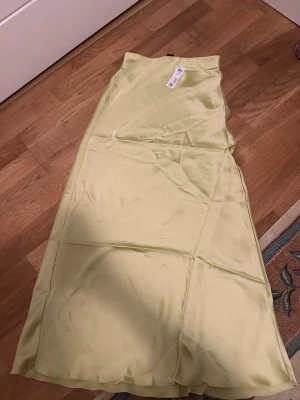 Ljusgul långkjol i satin - Säljer en ljusgul långkjol i satin med slät och glansig yta. Kjolen har en enkel, rak passform och är perfekt för dig som gillar stilrena plagg. Storlek XS enligt lappen.