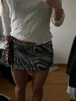 Zebramönstrad kortkjol med snörning - Säljer en kort zebramönstrad kjol i svart och silver med snörning i sidan. Kjolen har en tight passform och glittriga detaljer som gör den extra snygg.
