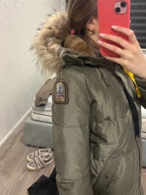 Olivgrön dunjacka från Parajumpers - Säljer en olivgrön dunjacka från Parajumpers med päls på huvan och gul detalj vid dragkedjan. Jackan har två snedställda fickor med knappar och ett snyggt märke på ärmen. Perfekt för kalla vinterdagar och riktigt varm.