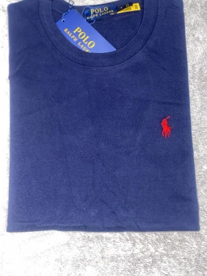 Marinblå t-shirt från Polo Ralph Lauren - Snygg marinblå t-shirt från Polo Ralph Lauren med klassisk röd broderad logga på bröstet. Tillverkad i mjuk bomull och har en enkel, stilren design med rund hals. Perfekt för dig som gillar tidlös och clean stil.