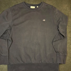 Mörkgrå GANT Sweatshirt / Crewneck (Strl M) - Säljer en klassisk och bekväm crewneck-tröja från GANT i storlek Medium. Tröjan har en snygg, naturligt urtvättad mörkgrå/svart ton som ger en skön vintagekänsla. Klassisk GANT-sköld broderad på bröstet. Skick: Använd med en schysst "washed" look och viss blekning vid sömmarna. Obs: Det finns några små fläckar på plagget. Titta noga på bilderna för att göra din egen bedömning av skicket. Säljes i befintligt skick enligt bilder. Storlek: M (Medium) Färg: Mörkgrå / Faded black Hör av dig om du har