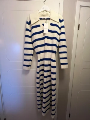 NY Randig stickad långklänning Adoore  - Säljer en lång ny stickad klänning från Adoore med vita och blå ränder. Klänningen har v-ringning, stor krage och långa ärmar. Materialet är ribbstickat och ger en mjuk och bekväm känsla. Perfekt för dig som gillar klassisk marin stil.