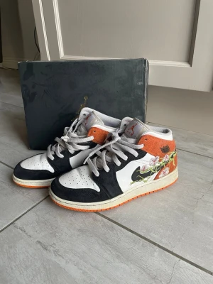 Nike Air Jordan 1 Orange/Blommig - Snygga Air Jordan 1  med svart, vitt och orange mocka och läder. Unik blommig design på sidan och klassisk Nike swoosh. Skön passform och ikonisk siluett som sticker ut. Perfekta för dig som vill ha något extra till din outfit. Vid frågor eller funderingar skriv gärna