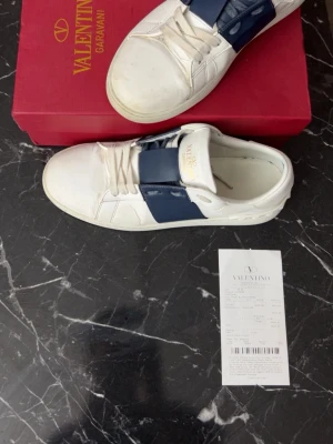Valentino Garavani Open sneakers vit/blå - Säljer ett par Valentino Garavani Open sneakers i vitt skinn med bred blå läderdetalj över snörningen. Skorna har rund tå, platt sula med mönster och ikoniska nitar baktill. Märkeslogga på plösen i guld. Snygg och stilren design, för fler frågor o bilder va inte tveksamma o priset går att diskutera 🤝