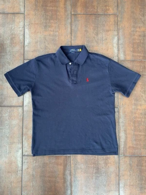 Marinblå pikétröja Polo Ralph Lauren  - Klassisk marinblå pikétröja från Polo Ralph Lauren med korta ärmar och krage. Tröjan har två knappar vid halsen och den ikoniska röda logotypen broderad på bröstet. Priset är ej hugget i sten, så kom med förslag! 