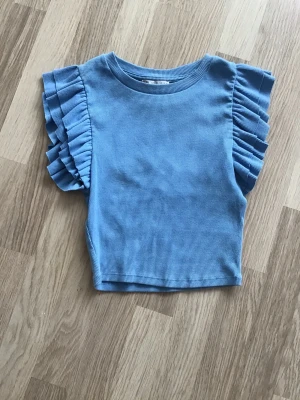 Blå ribbad topp med volangärmar från Zara, stl S - Supersöt blå topp med ribbad struktur och volangdetaljer på ärmarna. Croppad, kort modell som passar perfekt till både jeans och kjol. Just nu fri frakt, så du betalar bara för toppen. Jag har fler plagg ute, och hittar du något mer från min garderobsutrensning, så skickar jag allt i samma paket.