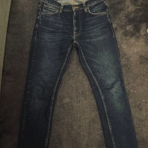 Mörkblå jeans från Nudie Jeans - Säljer ett par mörkblå jeans från Nudie Jeans med klassisk femficksmodell och kontrastsömmar. Jeansen har raka ben och snygga slitningar framtill samt Nudie Jeans ikoniska broderi på bakfickorna. Perfekta till en avslappnad stil. Skriv vid minsta fundering😁🔥