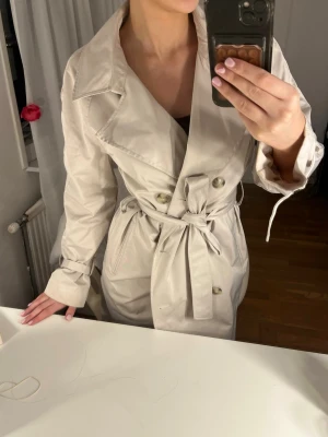 Beige trenchcoat med knytskärp - Stilren beige trenchcoat med dubbelknäppning och brett slag. Knytskärp i midjan och klassiska detaljer som axelklaffar och sidofickor. Lång modell som passar perfekt till lager på lager. Okänt märke och material.