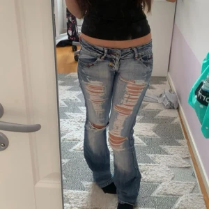 Lågmidjade crazy age jeans - Säljer ett par blå crazy age lågmidjade bootcut jeans med kraftiga slitningar och hål på framsidan av benen. Jeansen har låg midja, klassisk femficksmodell och ljus tvätt. Midja 36 cm innerben 80. De är igenlappade och ihop sydda på några ställen på rumpan för de har tillkommit tidigare hål(kan skicka bild på insidan). 