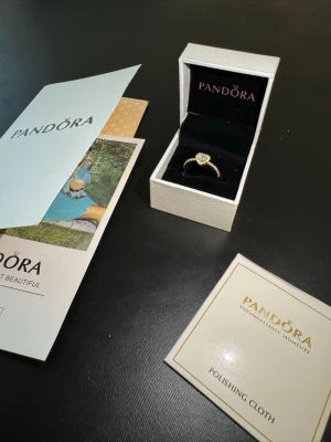 Guldfärgad hjärtring från Pandora - Säljer en elegant guldfärgad ring från Pandora med ett hjärtformat motiv och gnistrande stenar. Ringen har en detaljrik design med små stenar längs hela bandet och kommer i originalask med tillhörande putsduk och broschyr.