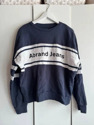Mörkblå sweatshirt från Abrand Jeans - Cool mörkblå sweatshirt från Abrand Jeans med vita ränder och logga framtill. Storlek S. Tröjan har rund hals, långa ärmar, breda muddar och nedhasade axlar. Mjuk o superskön kvalitét. Perfekt för en avslappnad och trendig look.  Använd 1 gång. Skrynklig på bilderna men den är i jättefint skick. 