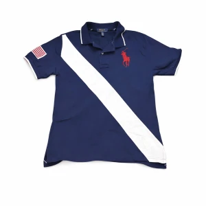 Marinblå pikéskjorta från Polo Ralph Lauren - Snygg marinblå pikéskjorta från Polo Ralph Lauren med vit diagonal rand framtill och röd logga på bröstet. Kortärmad modell med krage och knappar samt amerikansk flagga på ärmen. Perfekt för en sportig och stilren look. Den är vintage och säljs ej längre. Pikén är i bra skick och priset är ej hugget i sten och kan sänkas vid snabb affär!