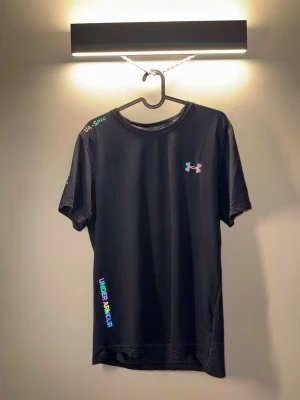 Svart träningsset från Under Armour - Snyggt svart träningsset från Under Armour med t-shirt och shorts. Båda plaggen har färgglada loggor och textdetaljer. Tillverkade i lätt och snabbtorkande material, perfekt för gymmet eller löprundan. T-shirten har rund hals och shortsen har resår i midjan.