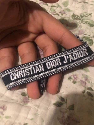 Christian Dior J'Adior armband - Snyggt armband från Christian Dior med texten 'J'Adior' broderat i vitt på en mörkblå bakgrund. Armbandet har dekorativa kanter och tofsar i ändarna, vilket ger en trendig och exklusiv känsla. Perfekt accessoar för att lyfta din outfit.