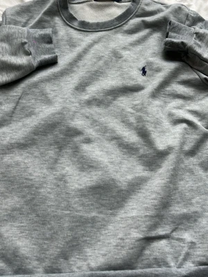 Grå sweatshirt från Ralph Lauren - Klassisk grå sweatshirt från Ralph Lauren med rund halsringning och broderad marinblå logga på bröstet. Tröjan har ribbade muddar vid ärmslut och nederkant. Perfekt för en avslappnad och stilren look.