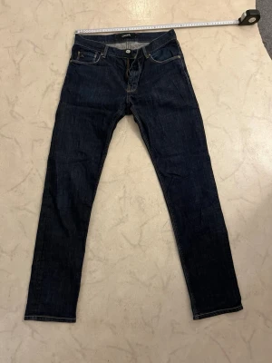 Mörkblå jeans från J.Lindeberg - 31/32 - Snygga mörkblå jeans från J.Lindeberg med klassisk femficksmodell och raka ben. Jeansen har kontrastsömmar och är tillverkade i slitstark denim. Perfekta för dig som gillar stilrena och tidlösa jeans.