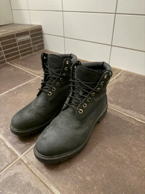 Timberland kängor  - Stilrena svarta kängor med grov sula och snörning. Kängorna har metallöljetter och vadderad krage för extra komfort. Tillverkade i ett matt, slitstarkt material som ger en klassisk look. Perfekta för dig som vill ha en robust och tidlös sko.