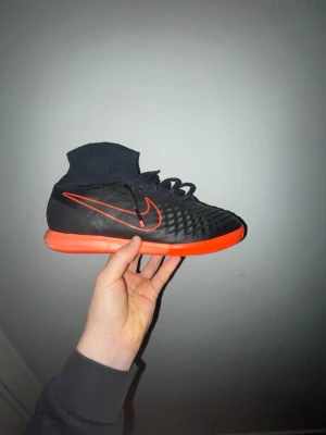 Nike svart/röd fotbollssko med strumpa - Säljer en svart Nike Magistax fotbollsskor med röd sula och röd swoosh. Skon har en stickad strumpliknande krage för extra stöd runt ankeln och snörning framtill. Ovandelen är texturerad för bättre bollkontroll och skon är designad för inomhusspel. Storlek 42