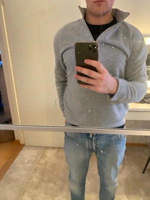 Grå halvzip tröja med hög krage - Säljer en stilren grå hoodie med halvzip och hög krage. Hoodien har långa ärmar och är tillverkad i ett mjukt material som passar perfekt för en avslappnad look. Enkel design utan synligt märke, perfekt att matcha med jeans. Storlek M