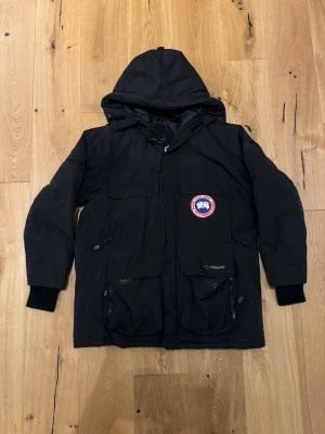 Svart dunjacka från Canada Goose - Svart dunjacka från Canada Goose med huva, flera stora fickor och klassisk logotyp på bröstet. Jackan är tillverkad i slitstarkt material och har ribbade muddar vid ärmslut. Perfekt för kalla vinterdagar och riktigt varm.