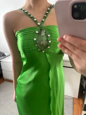 Grön klänning med strass och spetsdetalj - Snygg grön klänning med korsade axelband dekorerade med stora strass och en unik spetsdetalj framtill. Klänningen har en figurnära passform och är tillverkad i ett glansigt silkes material som ger en lyxig känsla. 