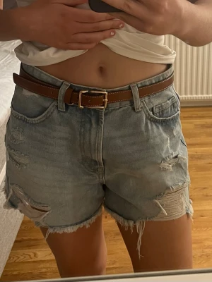Blå denimshorts med bälte - Säljer ett par ljusblå lågmidjade denimshorts med slitna detaljer och råa kanter. Shortsen har klassiska fickor fram och bak. Perfekta för sommaren! 