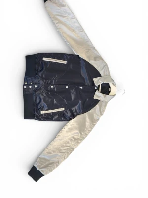 J Lindeberg jacka - Snygg bomberjacka i svart och silver med glansigt material. Jackan har vita detaljer vid fickorna, ribbade muddar och tryckknappar framtill. På ryggen finns ett färgglatt tryck med texten 'Thunderbirds Class of 1996' och en stadssilhuett. Perfekt för dig som vill sticka ut.