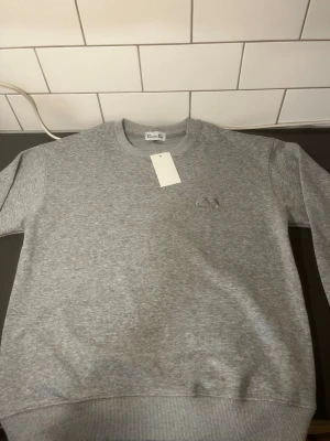CD tröja  - Snygg grå sweatshirt med en logga på bröstet. Klassisk rund halsringning och ribbade muddar vid ärmslut och nederkant. Perfekt för en clean och stilren look skickas inom 24 timmar passar för dig mellan 175-180cm