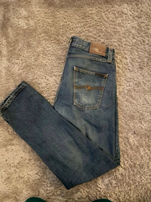 Blå jeans från Nudie Jeans - Säljer ett par klassiska blå jeans från Nudie Jeans med smal passform och snygga slitningar. Jeansen har fem fickor, orange sömmar och en läderpatch bak i midjan. Tillverkade i slitstarkt denim med en autentisk tvättad look.