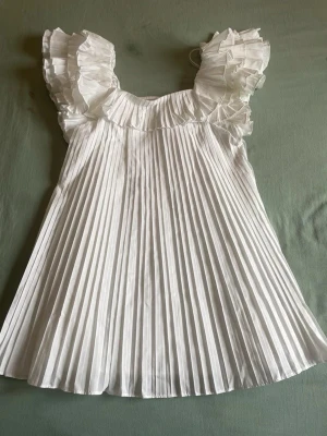 Vit plisserad klänning med volangärm - Supersöt vit klänning med plisserad kjol och stora volangärmar. Klänningen har en luftig och flowy passform med fyrkantig ringning. Perfekt för dig som gillar romantisk och trendig stil.