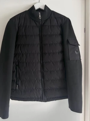 Svart pufferjacka från Moncler - Snygg svart pufferjacka från Moncler med quiltad front, ribbstickade ärmar och en ikonisk Moncler-logga på ärmfickan. Jackan har hög krage, dragkedja framtill och praktisk ficka med lock på ena ärmen. Perfekt för kalla vinterdagar. Passar M eller stor S, tror den är äkta, köpt på marketplace