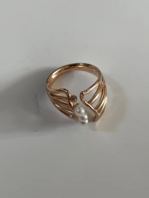 Guldfärgad ring med pärlor - Unik guldfärgad ring med ett öppet, vågigt mönster och två små vita pärlor framtill. Ringen har en elegant och modern design som sticker ut och passar perfekt för dig som gillar detaljer. Materialet ser ut som metall med en blank finish.