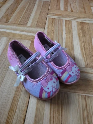 Hello Kitty ballerinaskor rosa - Säljer ett par söta rosa ballerinaskor från Sanrio med Hello Kitty-motiv och regnbågsdetaljer. Skorna har glittrande kant, vita rosetter på remmen och är tillverkade i textilmaterial. Perfekt för dig som gillar gulliga och färgglada skor.